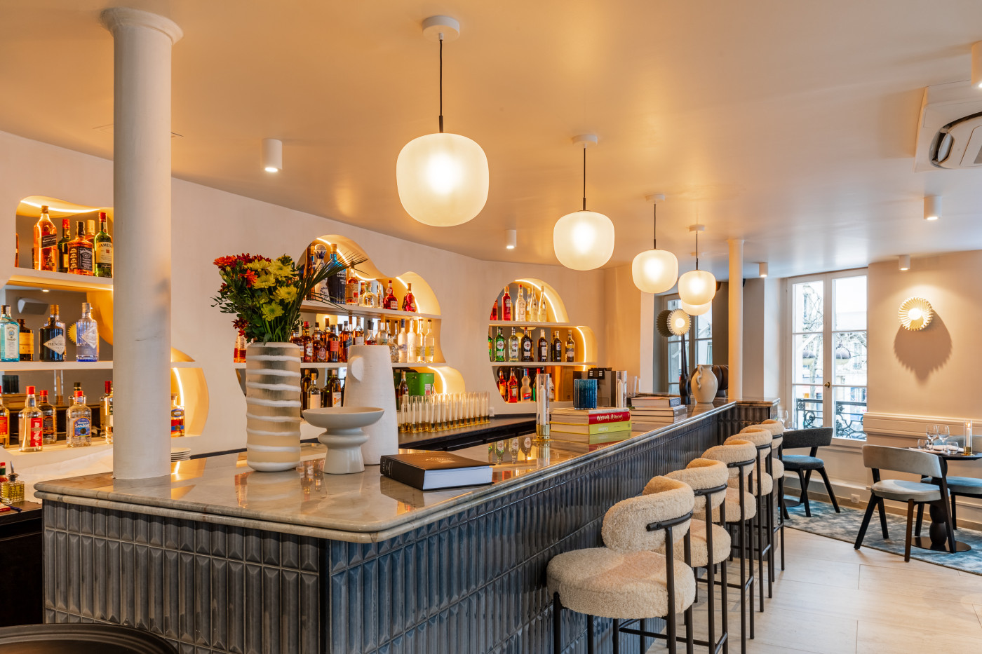 Le bar du Café des Ternes habillé de zellige bleu, ses tabourets en bouclette et ses suspensions en verre opalin
